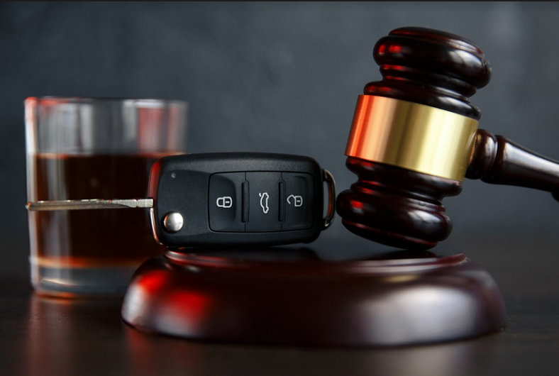 A wooden gavel and sound block with a car key and a glass of whiskey on a dark background.