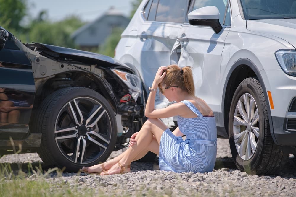 Distressed woman sitting by damaged cars after an accident, holding head in disbelief and worry.