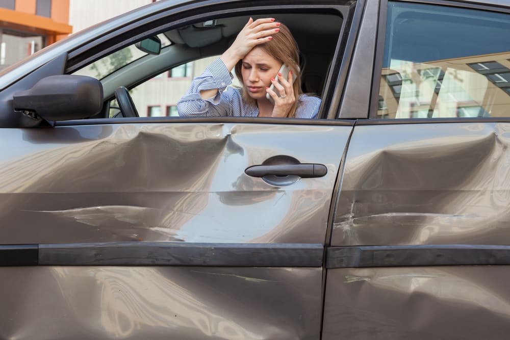 Woman in crashed car, phone in hand, wears a despairing expression, hand on forehead, conveying distress after road accident.