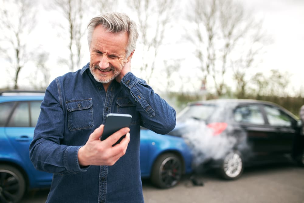 Senior driver with whiplash stands by wrecked car, reports accident to insurance via mobile phone.