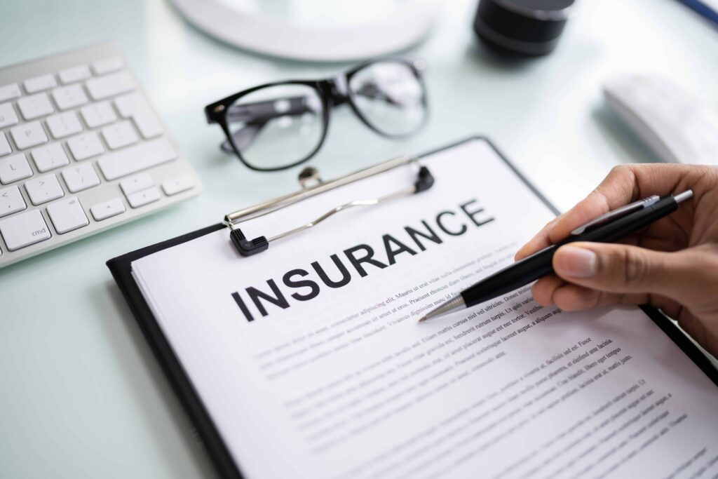 Person reviewing and signing an insurance document with pen on desk beside glasses and keyboard.